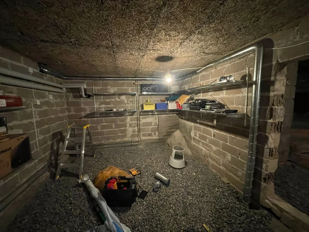 Grovkällare med grusgolv och oputsade tegelväggar i ett äldre hus, där radonsugens galvade kanal löper ner längs en pelare och vidare ned i grusbädden