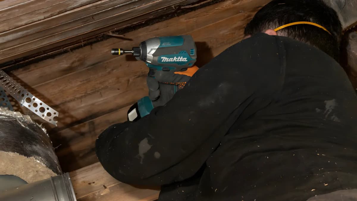 Tekniker sedd bakifrån fäster ett montageband i takträet med skruvdragare, en isolerad ventilationskanal syns intill