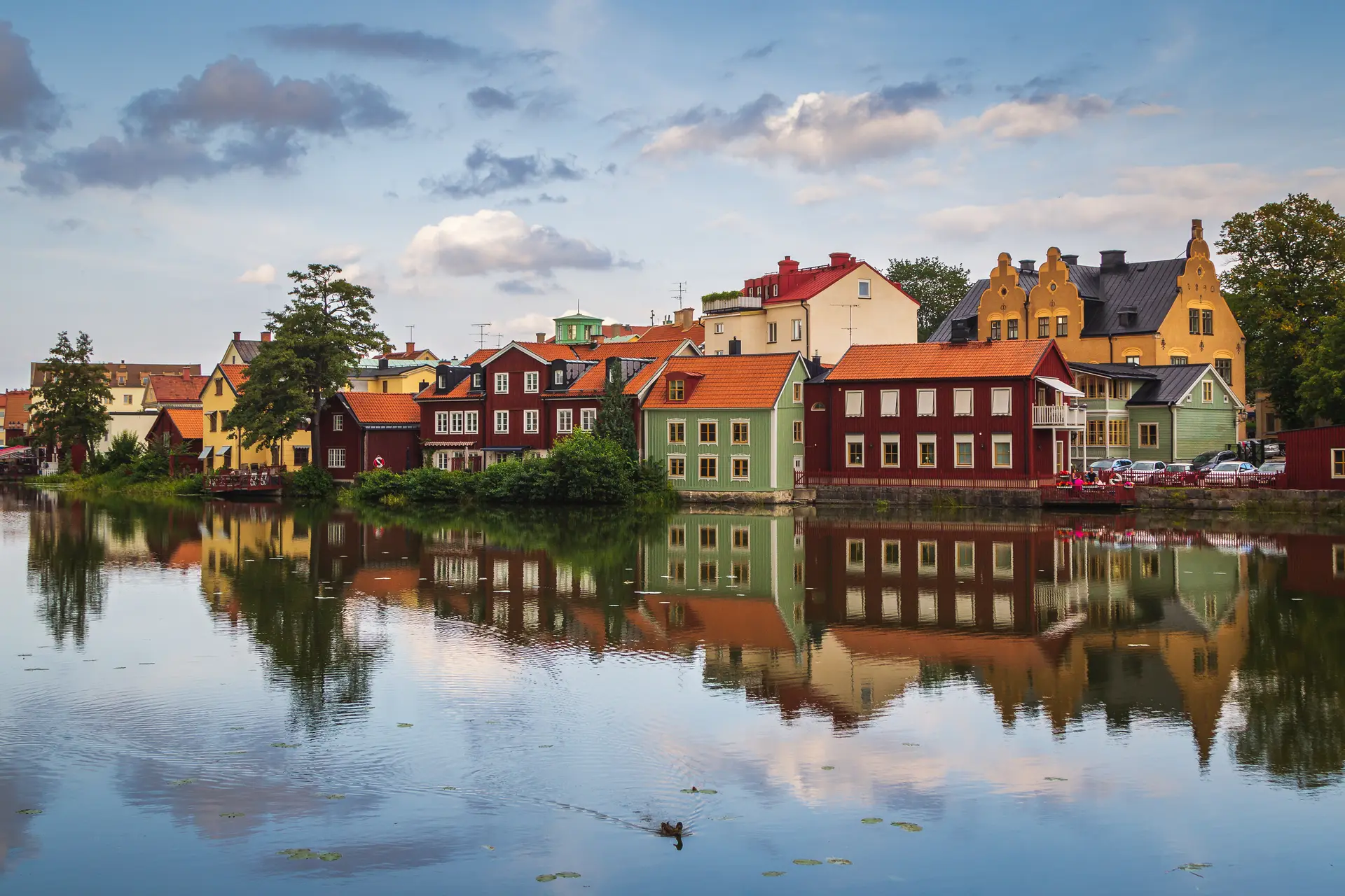 En and simmar förbi de rödmålade träfasaderna vid åstranden i Eskilstuna medan kvällssolen färgar molnen
